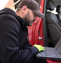 Mann in schwarzer Kleidung und Mütze, der an einem Laptop im Innenraum eines Autos arbeitet.
