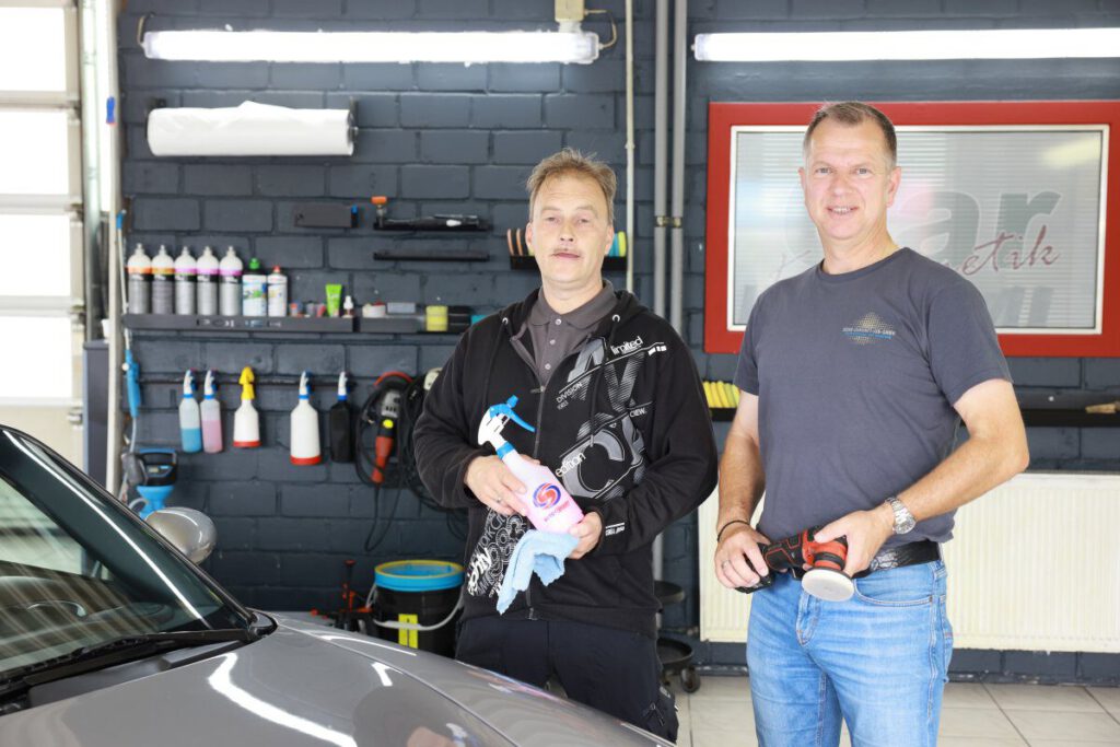 Zwei Männer halten Autopflegeprodukte in einer Werkstatt mit verschiedenen Reinigungsmitteln im Hintergrund.